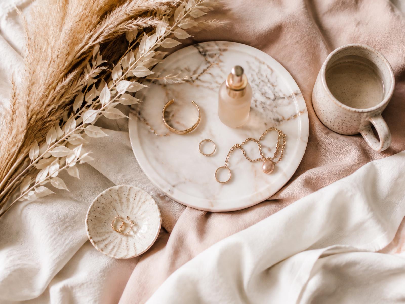 Flat lay of blush linen, gold jewelry, a ceramic mug and a skincare bottle